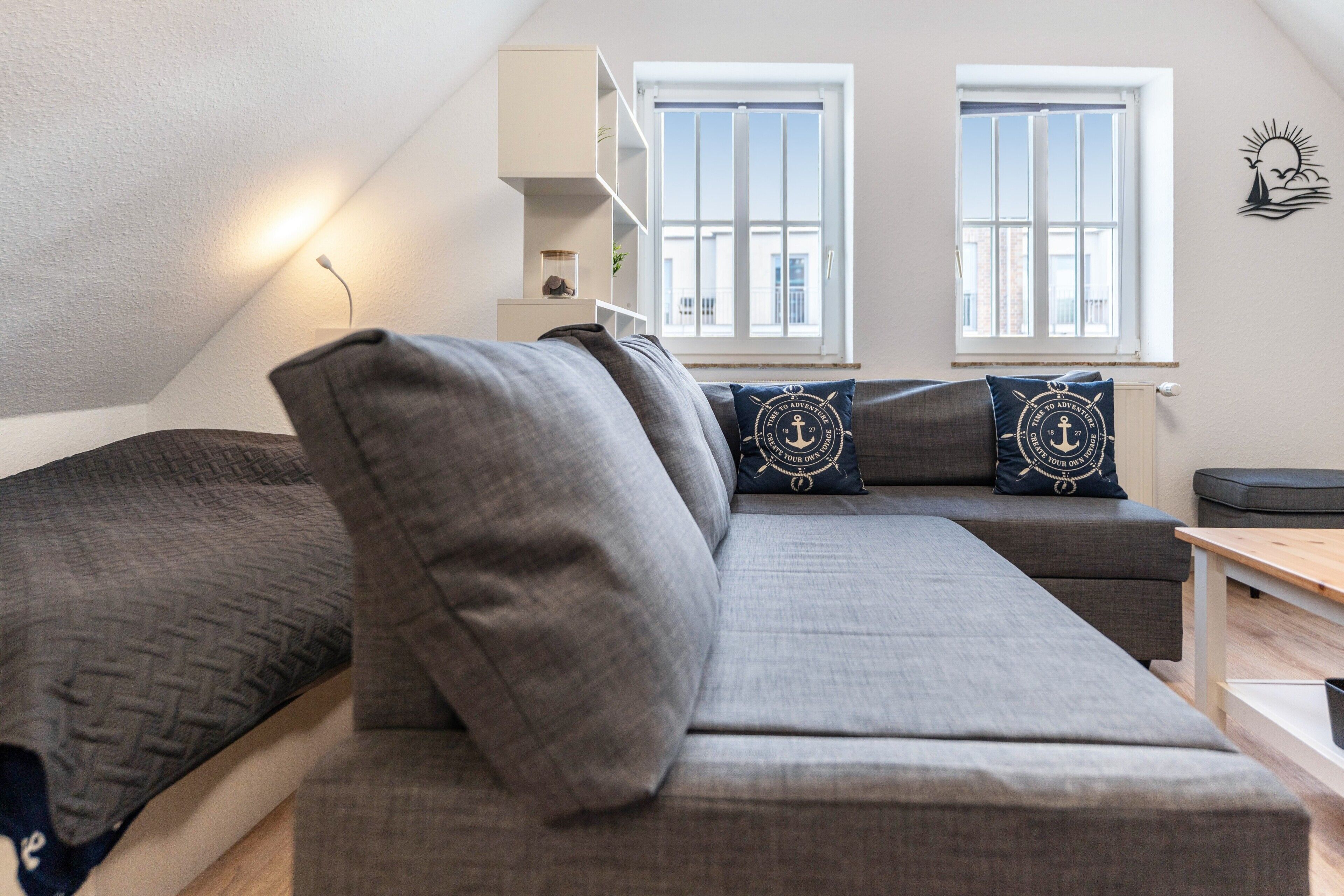Ein modernes Schlafzimmer im Loft-Stil mit einem großen grauen Sofa, zwei Fenstern mit weißen Rahmen und einem an der Wand montierten Regal.