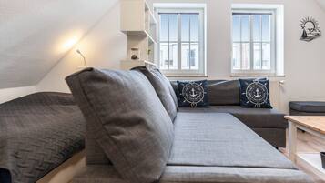 Ein modernes Schlafzimmer im Loft-Stil mit einem großen grauen Sofa, zwei Fenstern mit weißen Rahmen und einem an der Wand montierten Regal.