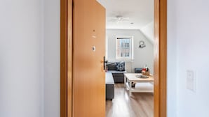 Een gang die uitkomt in een woonkamer met een bank, een salontafel en een raam met jaloezieën.