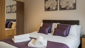 A neatly made bed with a purple bedspread, white pillows, and a folded white towel. There are two large floral paintings on the wall above the bed. - Westview Luxe Apartment (Newsome)