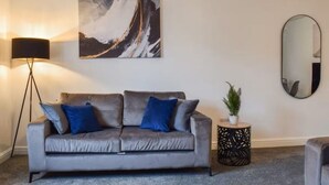 A living room with a grey sofa, blue cushions, a floor lamp, a round side table with a plant, a large abstract painting, and a mirror on the wall. - Westview Luxe Apartment (Newsome)