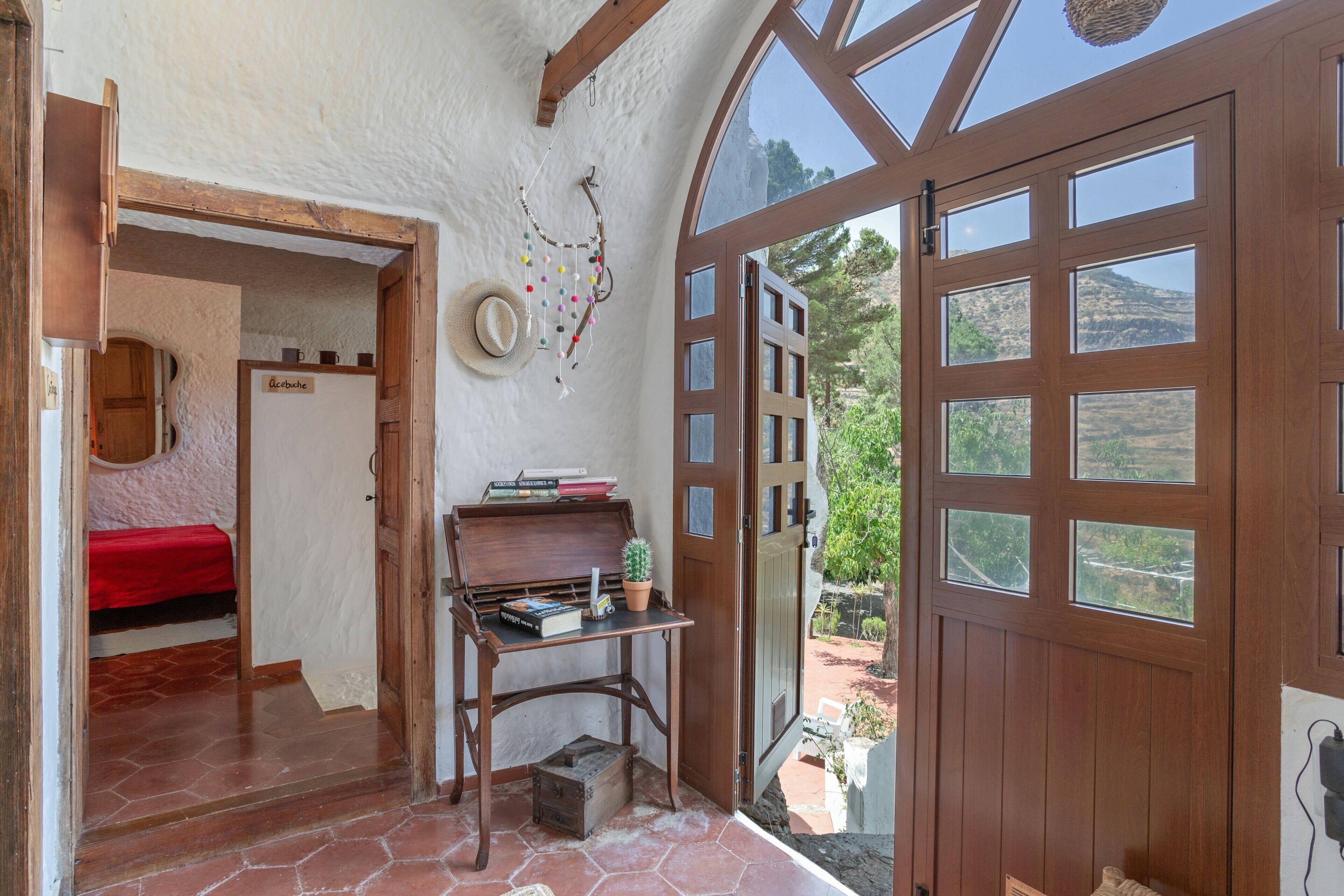 Una habitación con puerta de madera, una pequeña mesa con libros y una planta, cama roja y vistas al exterior.