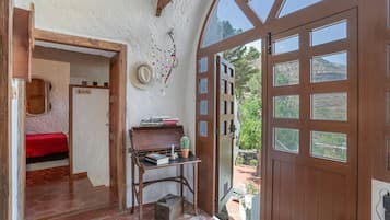 Una habitación con puerta de madera, una pequeña mesa con libros y una planta, cama roja y vistas al exterior.