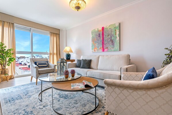 A modern living room with a sofa, armchairs, a coffee table, and a large window offering a city view.
