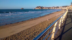 Ein Küstenweg mit Kiesstrand, Meereswellen und einer in der Ferne liegenden Stadt.
