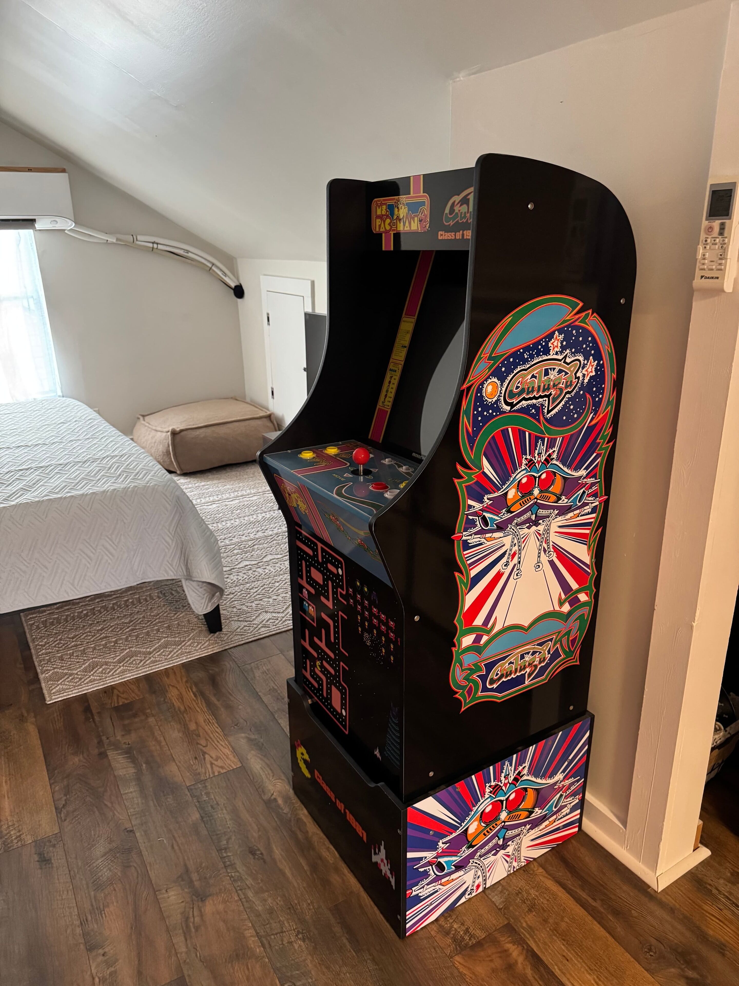 Une pièce avec un lit, une petite table et une borne d’arcade aux couleurs vives.