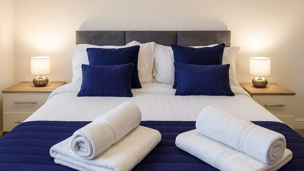 A neatly made bed with white and navy bedding, two bedside lamps, and rolled towels on the bed.