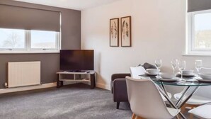 A modern living room with a flat-screen TV, a glass dining table set with plates and glasses, a radiator, and two framed artworks on the wall.