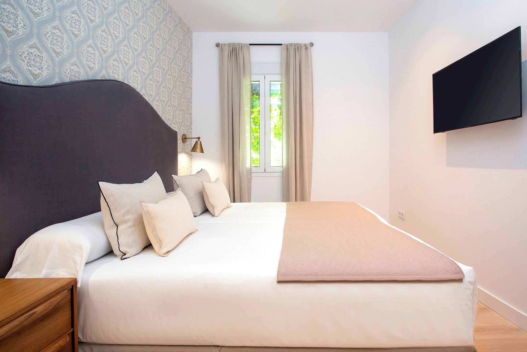 Un lit bien fait, avec une tête de lit, une table de chevet, une lampe et un téléviseur fixé au mur.