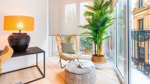 Een moderne woonkamer met een groot raam, een rieten stoel, een rond hocker, een zwart bijzettafeltje met een lamp en een potplant.