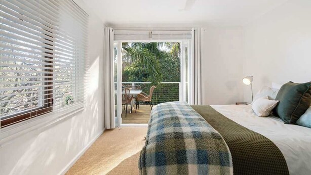 A bedroom with a bed, a bedside lamp, and a view of a balcony with outdoor furniture. - The Hidden Cottage (Tootgarook)