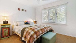 A bedroom with a bed, a bedside table with a lamp, a window with blinds, and a ceiling fan. - The Hidden Cottage (Tootgarook)
