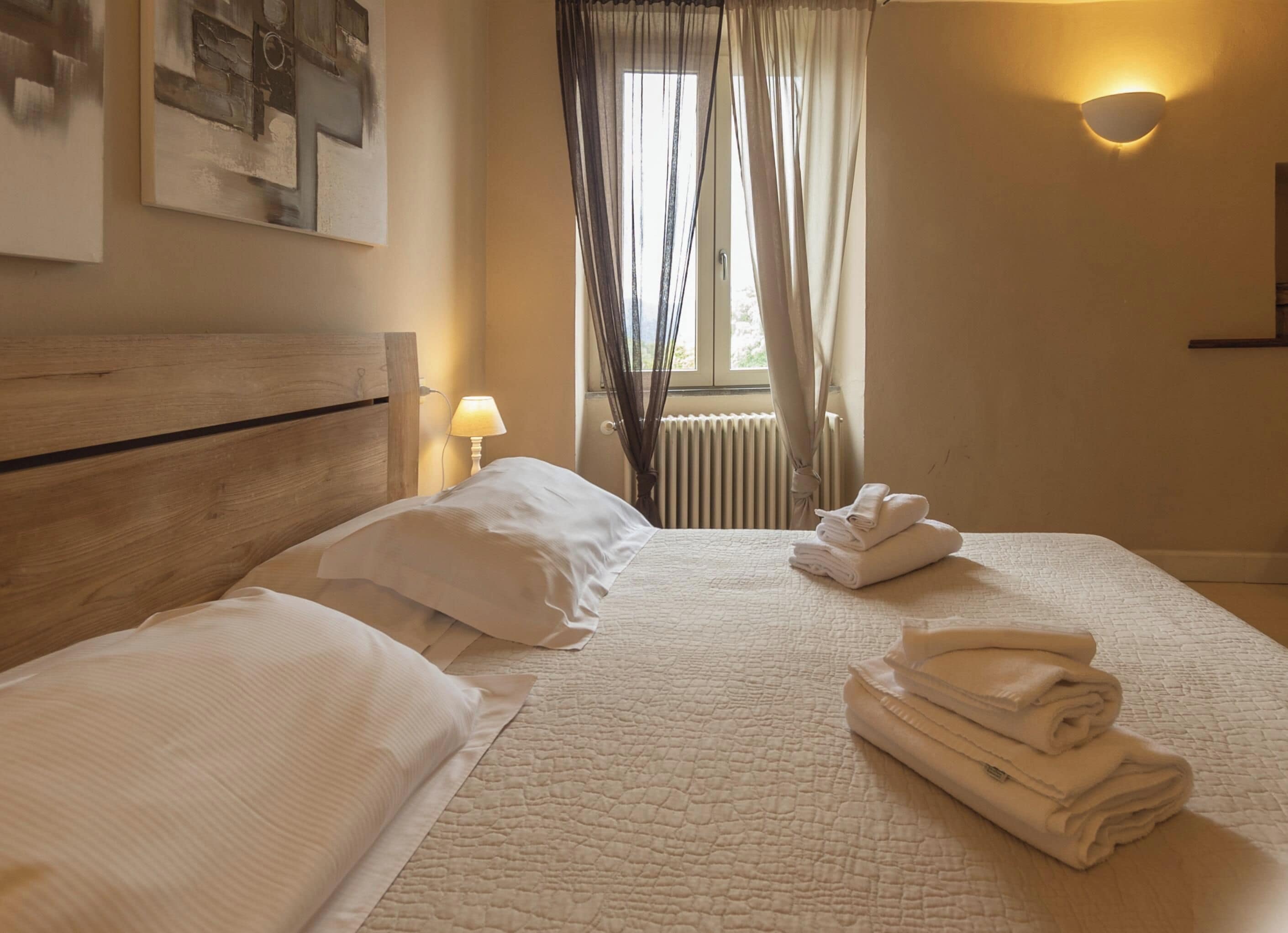 A hotel room with a bed, pillows, a folded towel stack, a wall-mounted lamp, and a window with curtains.