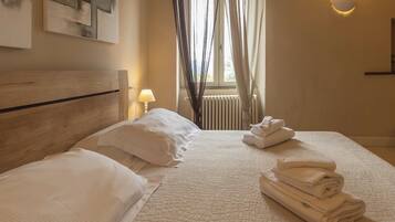 Habitación de hotel con una cama, almohadas, una pila de toallas dobladas, una lámpara de pared y una ventana con cortinas.