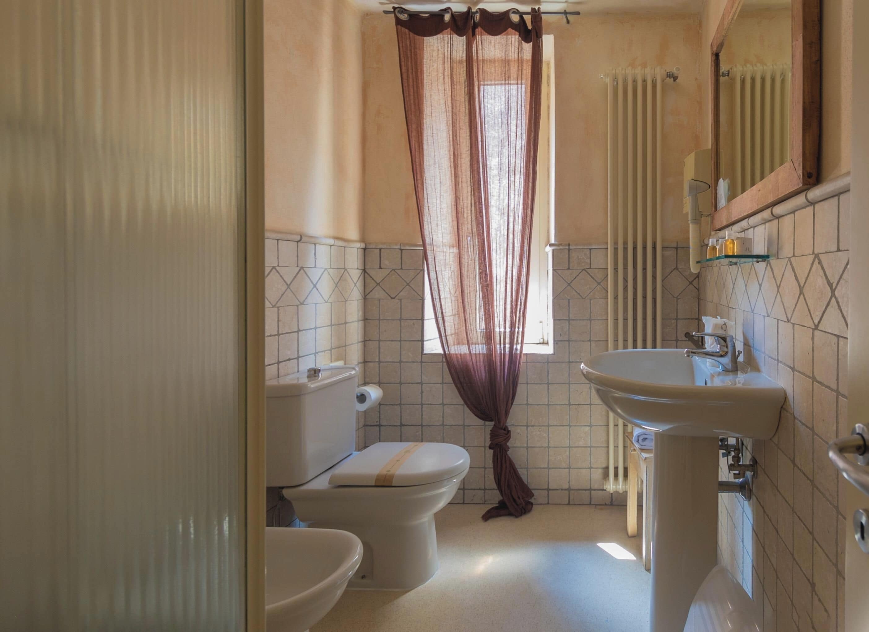 A bathroom with a toilet, bidet, and sink, a window with a curtain, and a mirror above the sink.