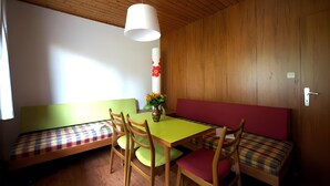 Ein Zimmer mit Holzboden, einem grünen Tisch, Holzsesseln, einer Bank mit kariertem Kissen und einer Vase mit Blumen.