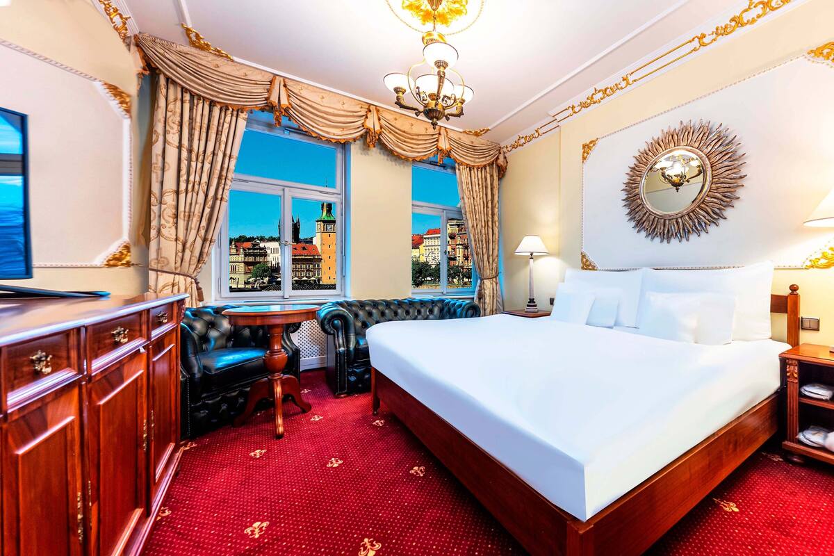 A luxurious hotel room with a large bed, a view of the city through a window, a chandelier, and a decorative mirror.