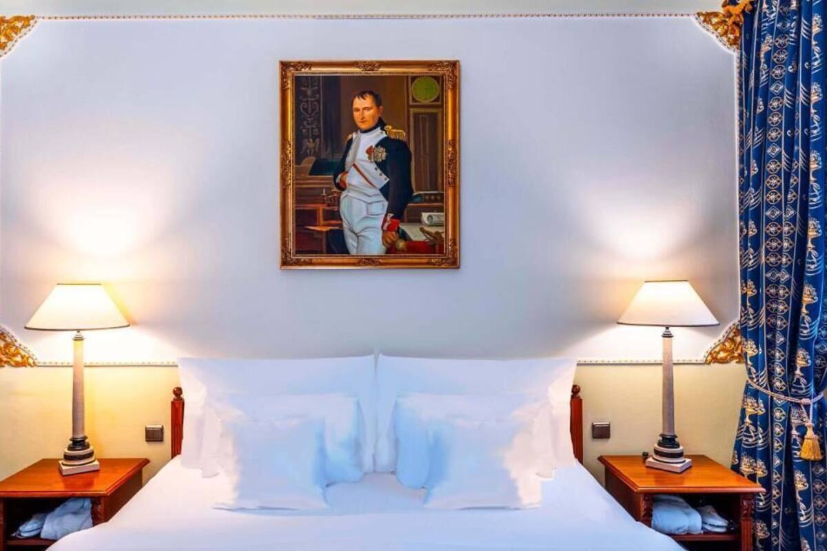 A hotel room with a large bed, two bedside tables with lamps, a framed portrait on the wall, and a patterned curtain.