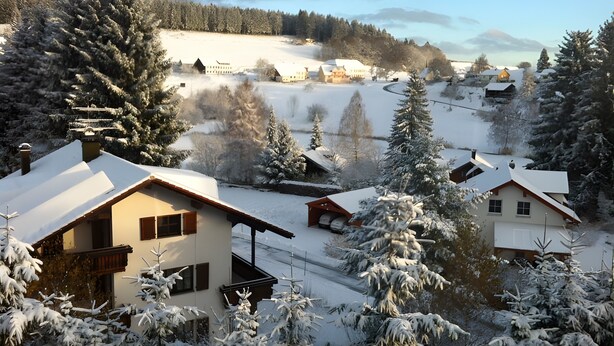 Un village enneigé, avec des maisons traditionnelles, un ciel dégagé et un lac en arrière-plan.