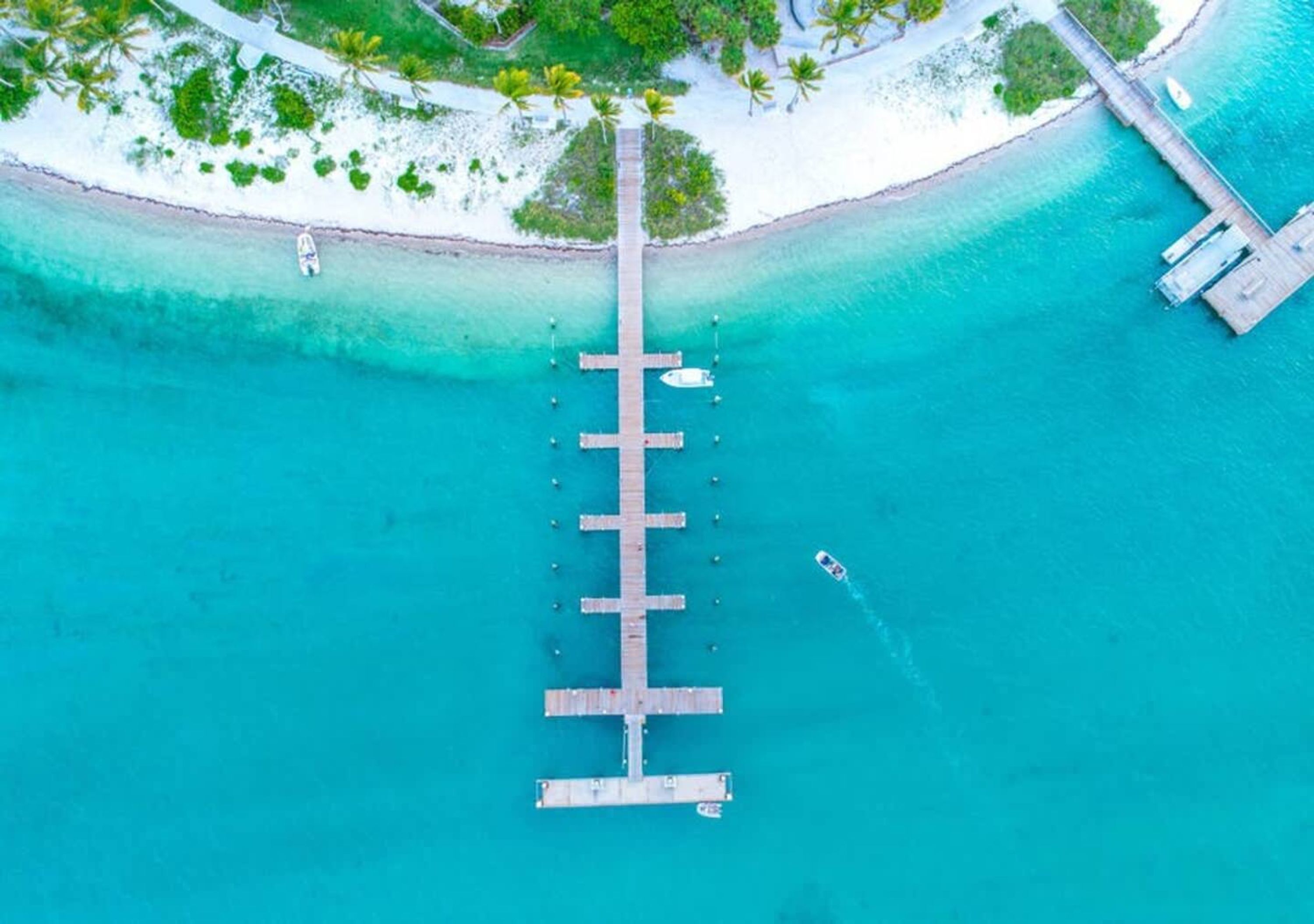 An aerial view of a pier extending into turquoise waters, surrounded by white sand and palm trees.