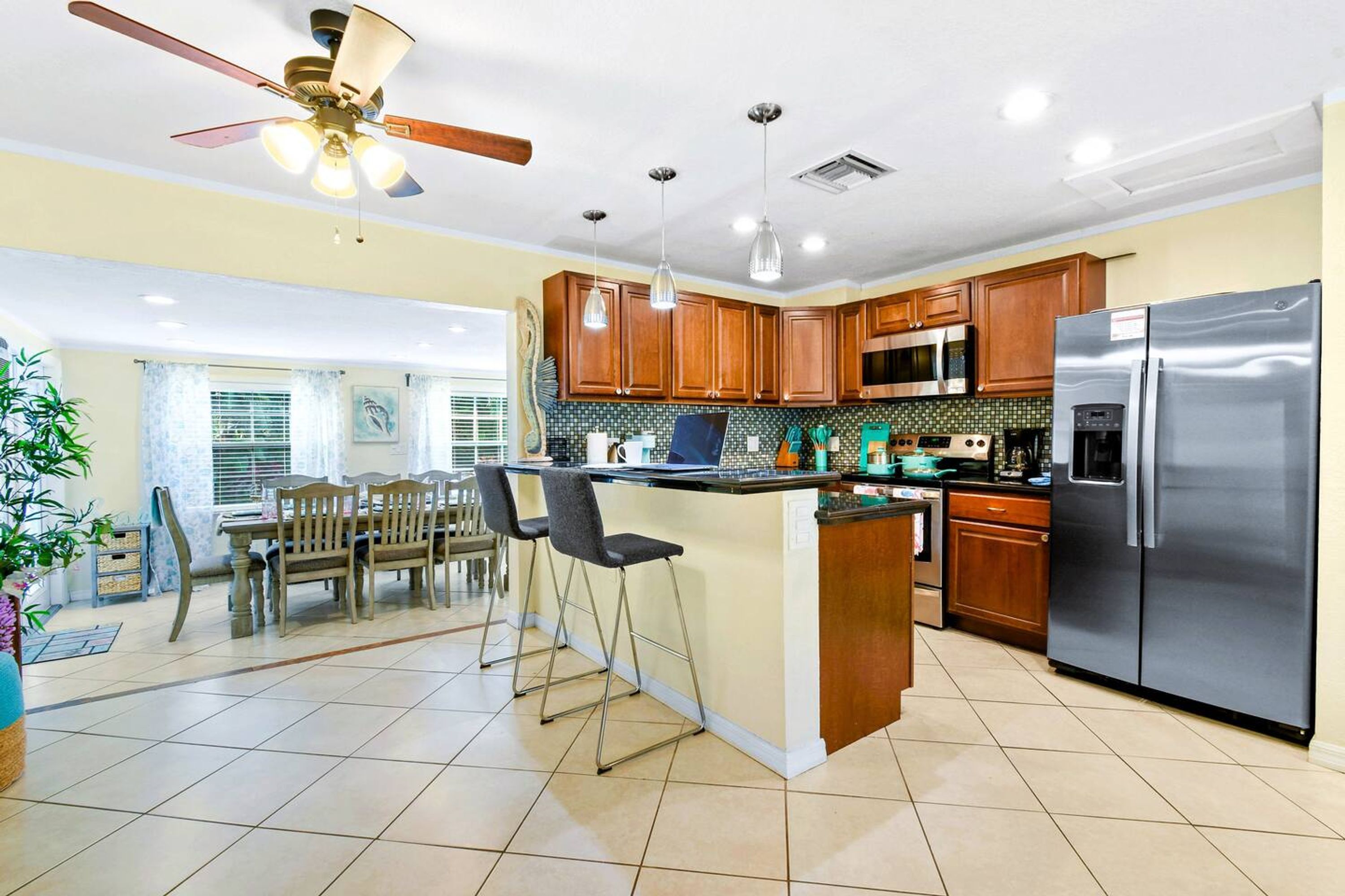 A modern kitchen with wooden cabinets, a stainless steel refrigerator, a dining area with a table and chairs, and a ceiling fan.
