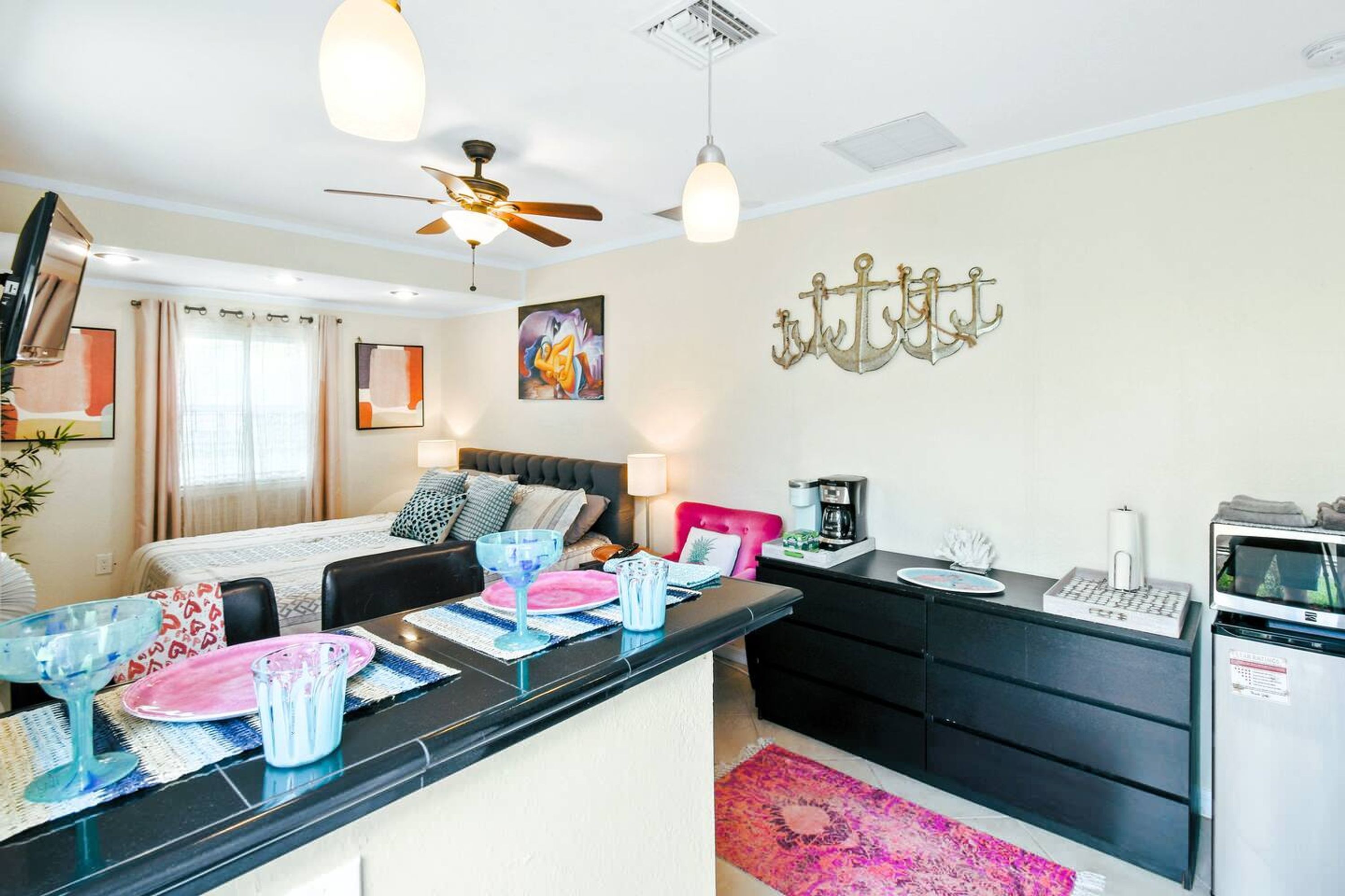 A modern hotel room with a bed, bedside table, desk, and kitchenette.