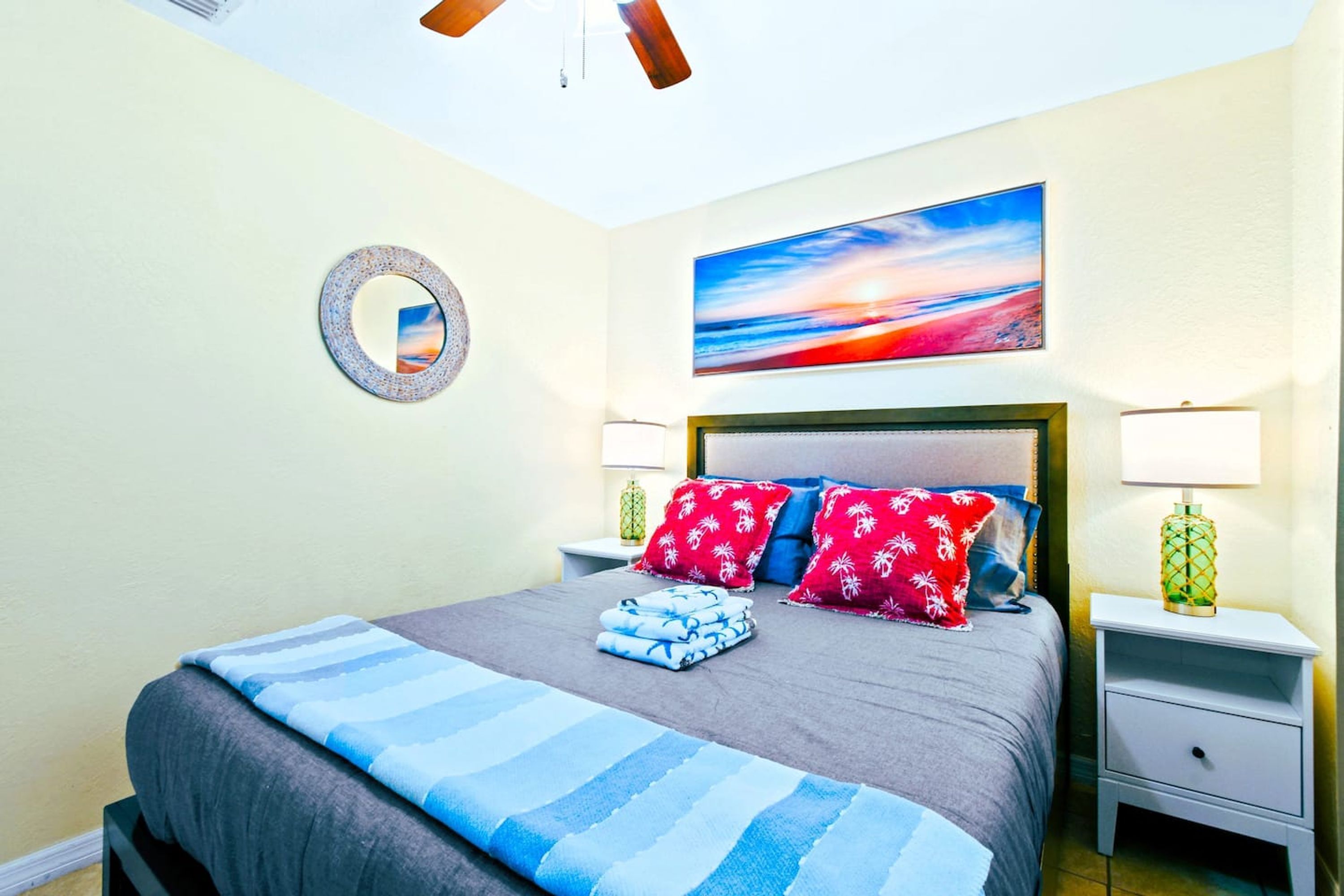 A neatly made bed with striped bedding, a headboard with decorative pillows, a wall-mounted mirror, a painting of a sunset, and two bedside tables with lamps.