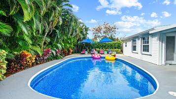 A swimming pool with a clear blue water surface, surrounded by lush greenery and a white building with windows.