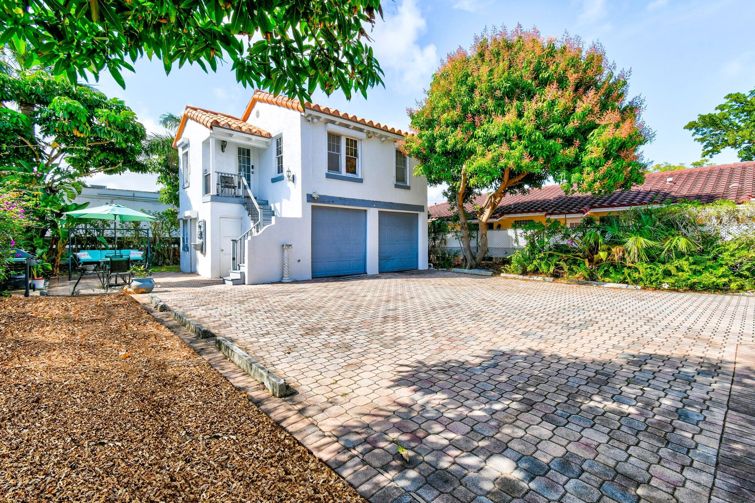 Une maison à deux étages, avec un toit en tuiles, un patio couvert aménagé avec du mobilier d’extérieur et une allée menant à un garage.