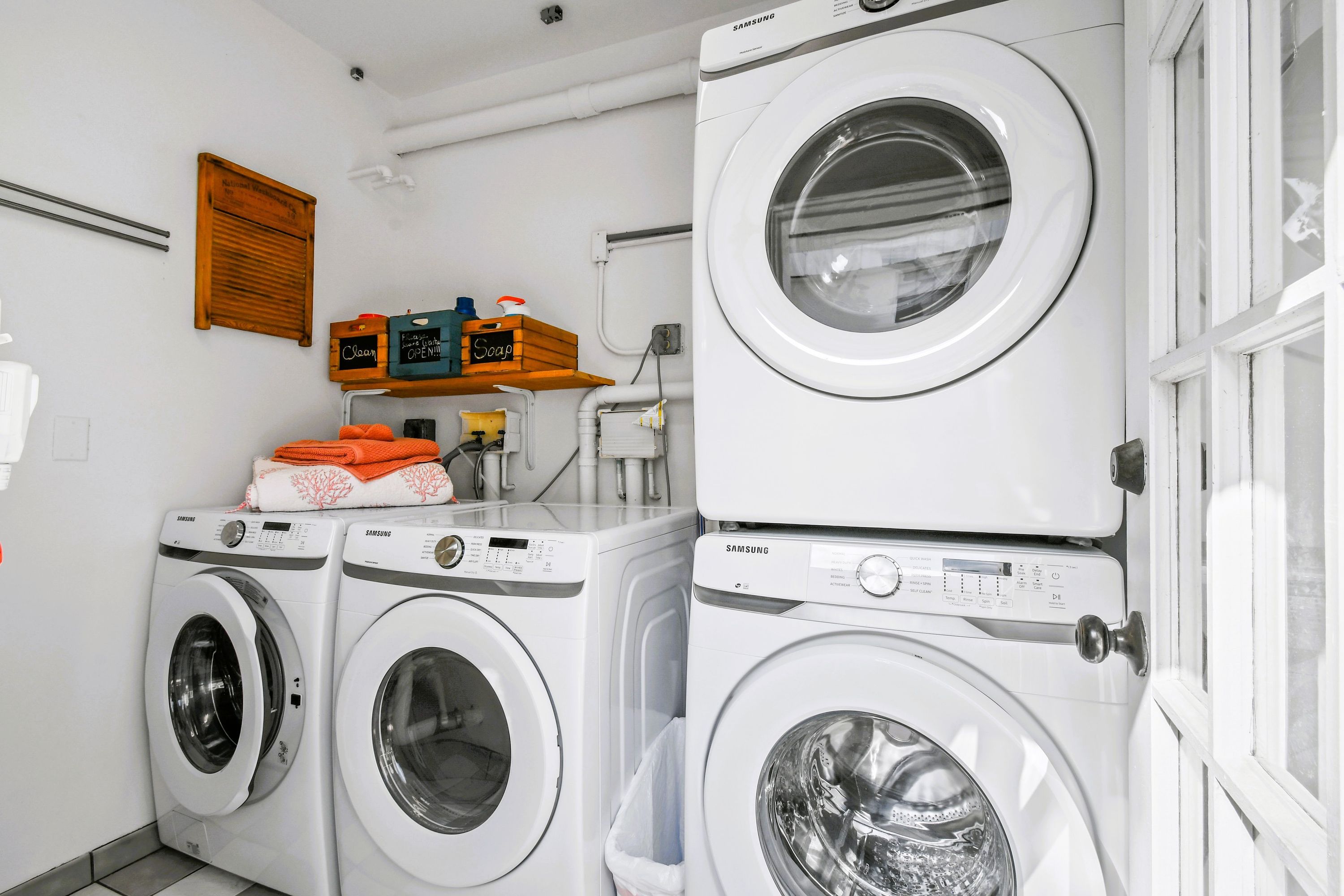 A laundry area with three stacked washing machines, a shelf with cleaning supplies, and a wooden sign on the wall.