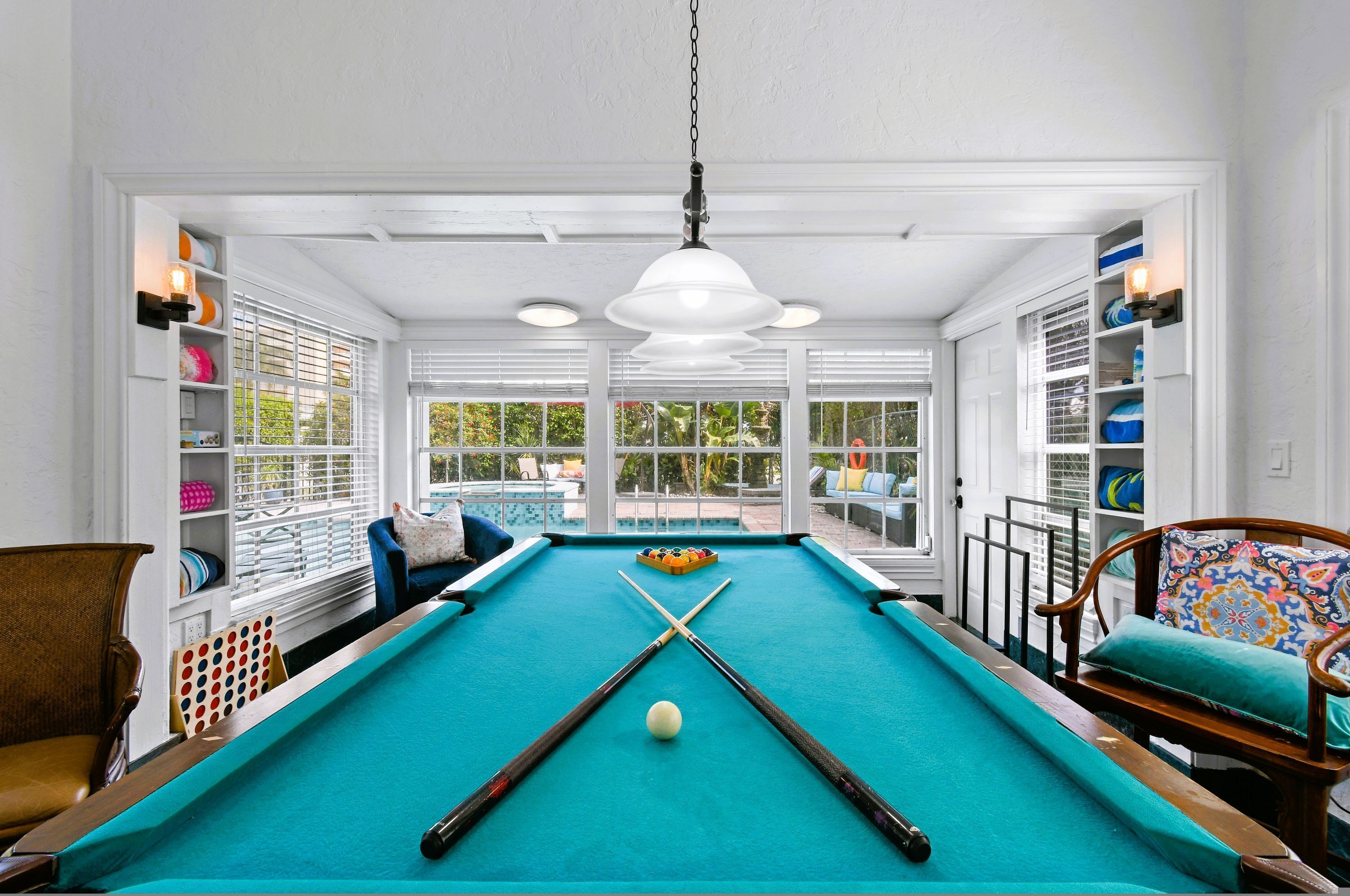 A pool table with a white ceiling, wooden chairs, and a view of a pool outside.