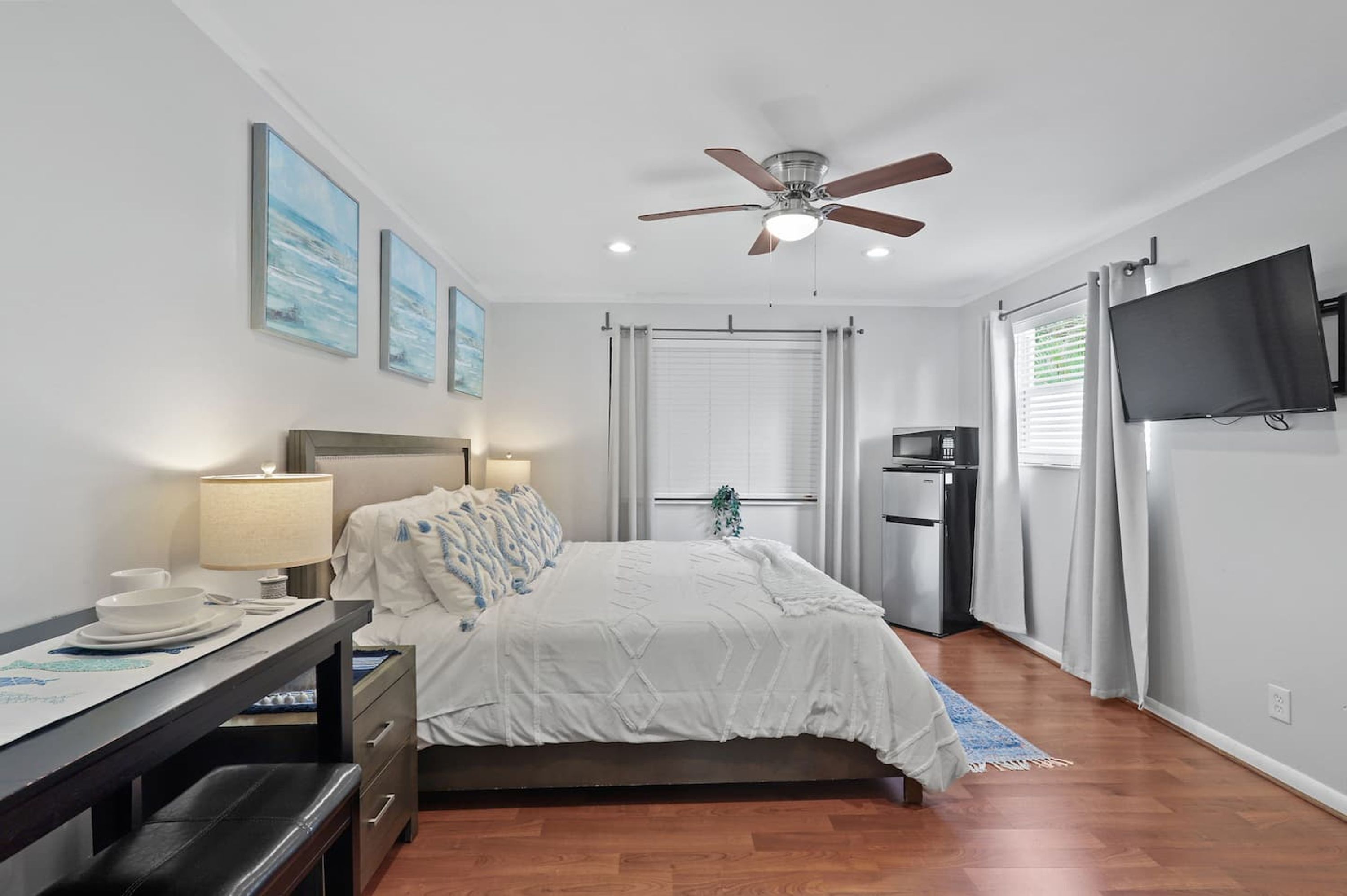 A bedroom with a bed, a desk with a lamp, a ceiling fan, a TV, and a window with curtains.