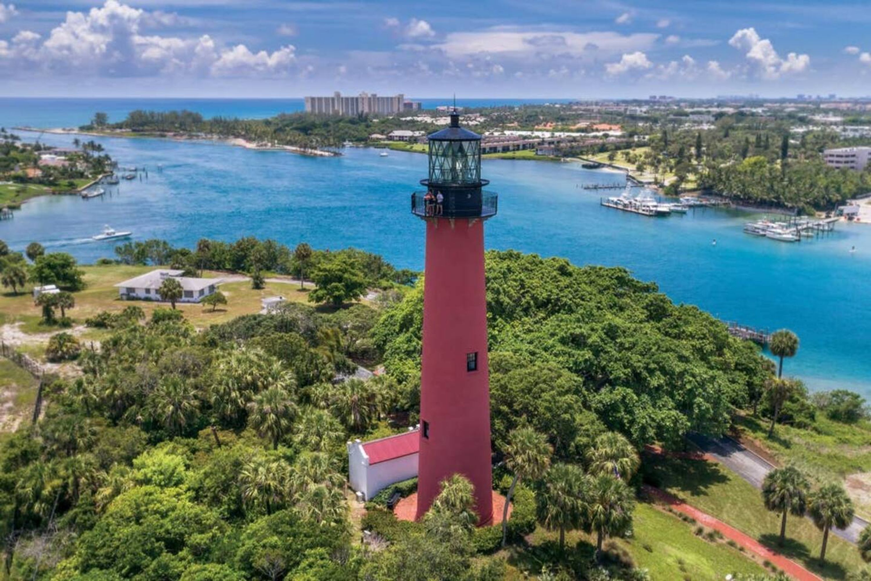 A red lighthouse surrounded by lush greenery and a marina with boats.
