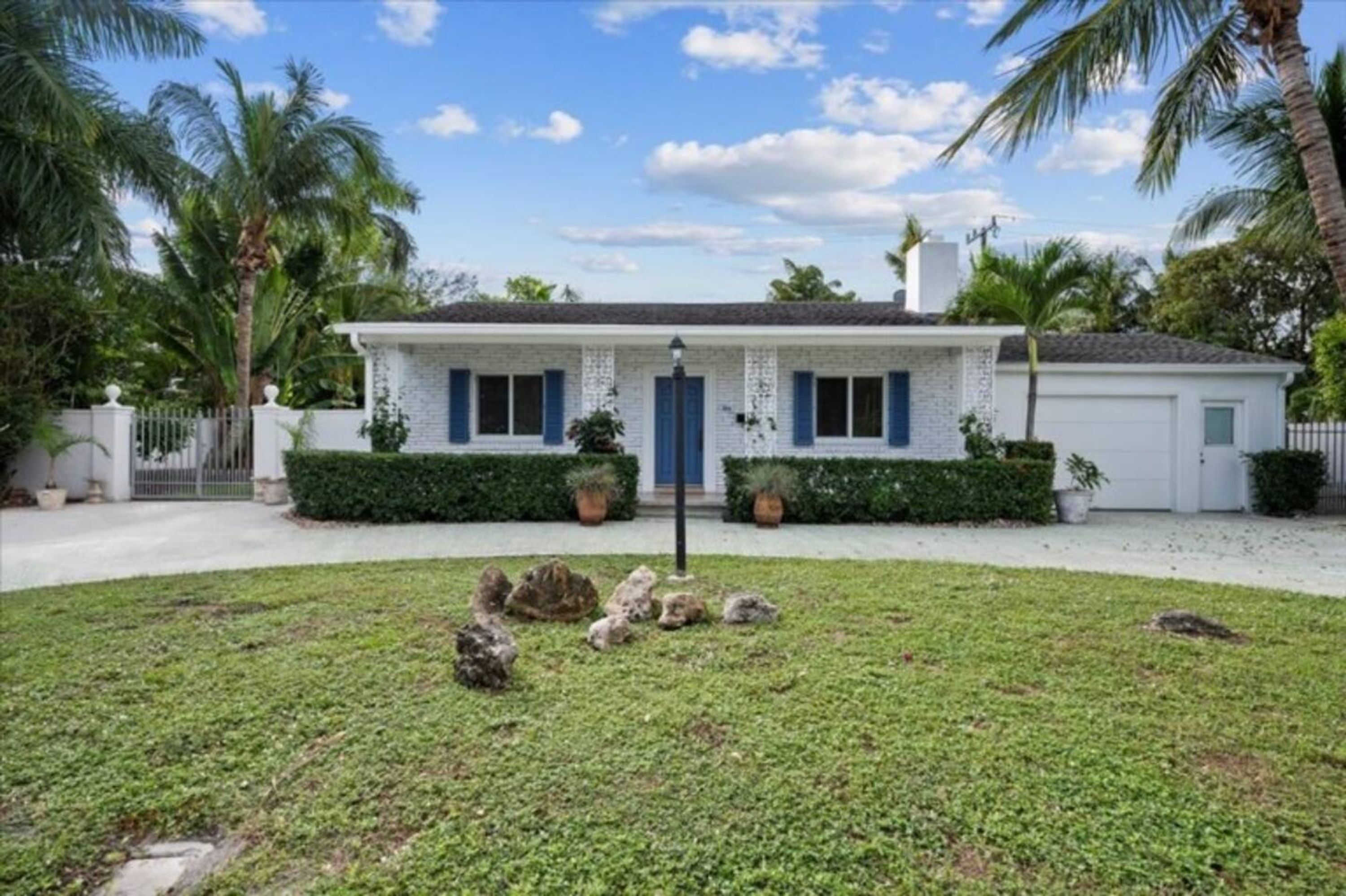 A single-story house with a white exterior and blue shutters, surrounded by a well-maintained lawn and palm trees.