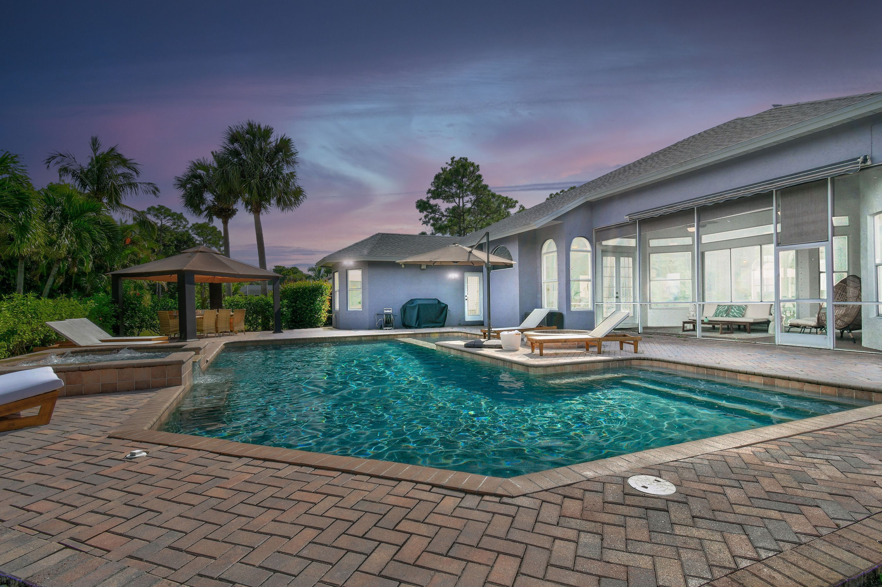 A swimming pool with lounge chairs and a gazebo, surrounded by palm trees and a house with large windows.