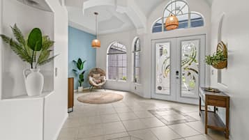 A spacious hallway with white walls, a tiled floor, and a large window. There are two wicker lampshades hanging from the ceiling, a wicker chair, and a small wooden table with a basket of plants.