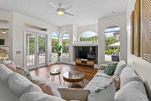 A spacious living room with a large sofa, a fireplace, and a television. There are two small tables and a ceiling fan. - Why Is Everyone Obsessed With This House (Loxahatchee)