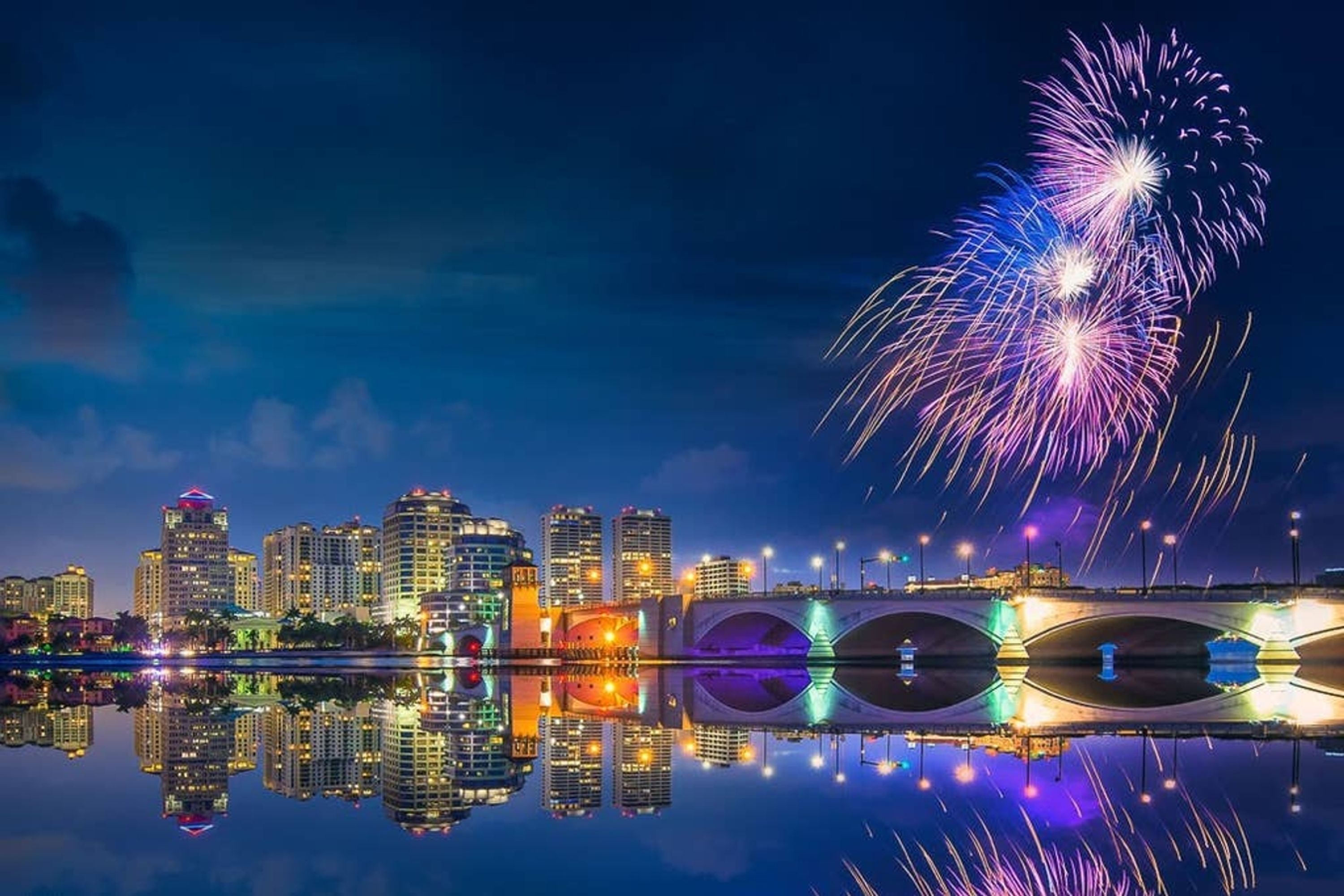 Un horizonte urbano con edificios iluminados reflejados en aguas tranquilas, un puente con luces y fuegos artificiales en el cielo.