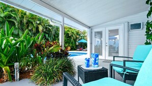 A covered outdoor area with a pool, surrounded by lush tropical plants and furnished with wicker chairs and a table. - NEW Home w Guest Suite Pool Near PGA Beach (Palm Beach Gardens)