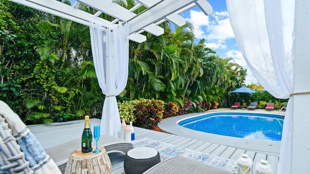 A poolside area with a wooden deck, lounge chairs, a small table with a bottle and glasses, and lush greenery in the background. - NEW Home w Guest Suite Pool Near PGA Beach (Palm Beach Gardens)