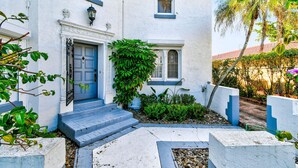 A white building with a blue door, a small garden with plants, and a paved pathway leading to the entrance. - 1926 Mediterranean Home With Heated Pool Sleeps 12 (West Palm Beach)