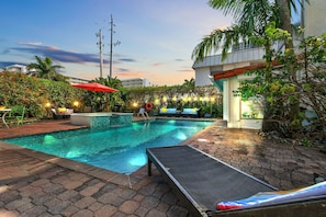Piscina com espreguiçadeira, guarda-sol vermelho e caminho de tijolos.