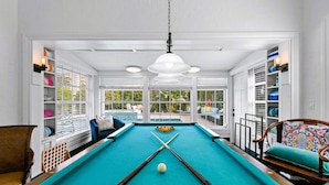 A pool table with a teal felt surface, surrounded by white walls and large windows. - 1926 Mediterranean Home With Heated Pool Sleeps 12 (West Palm Beach)