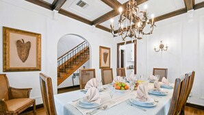 A dining room with a long table set for a meal, wooden chairs, a chandelier, and framed artwork on the walls. - 1926 Mediterranean Home With Heated Pool Sleeps 12 (West Palm Beach)