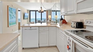 A modern kitchen with white cabinets, a dishwasher, a stove, and a large window overlooking the ocean. - Beachfront 2BR condo in Marco Island with pool and WiFi. (Marco Island)
