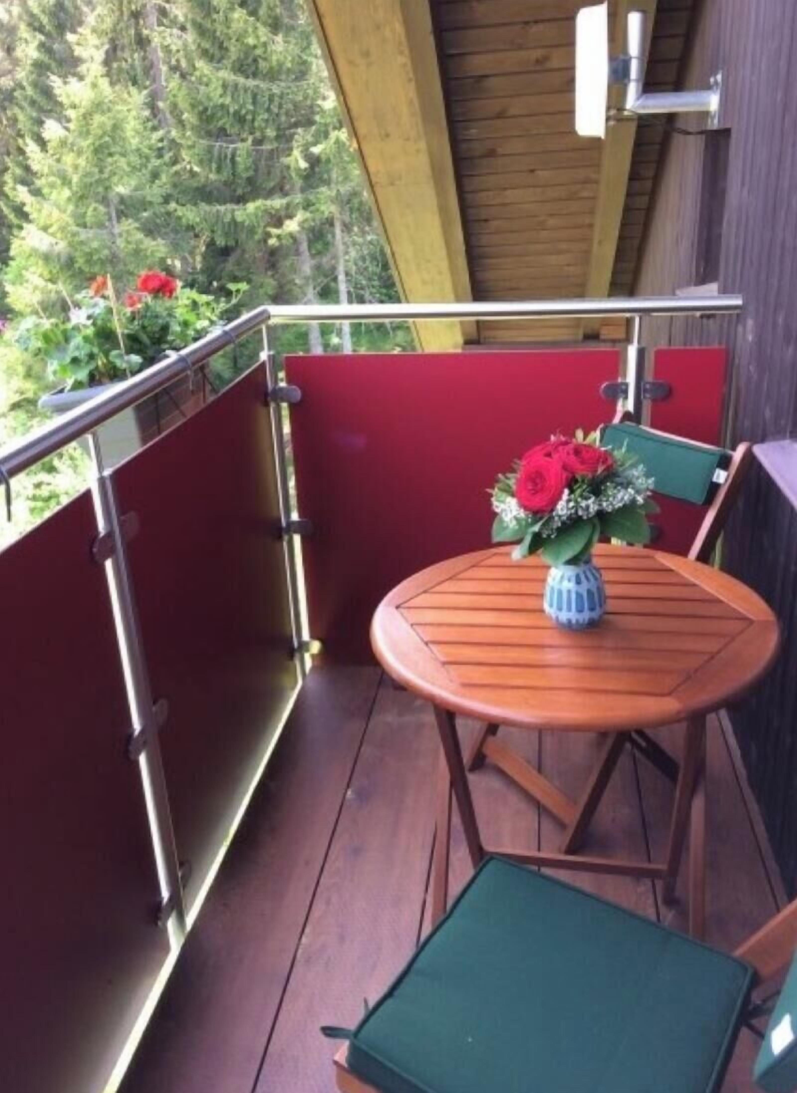 Una terraza de madera con una mesa redonda y sillas, un jarrón con flores y vistas al verdor.