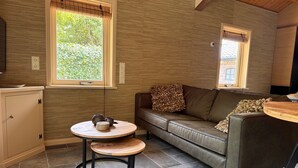 A cozy living area with a brown leather sofa, a round wooden table, and a small stool. There is a window with a view of greenery outside. - Chalet with privacy and garden near Leeuwarden (Jelsum)