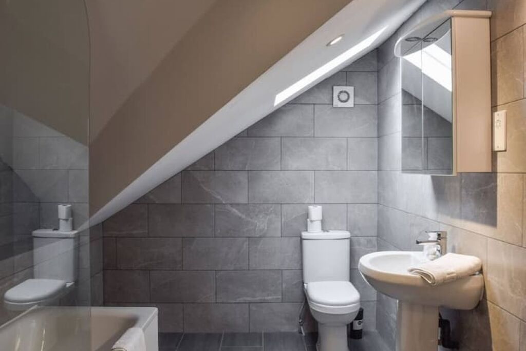 Et moderne badeværelse med skråvæg, grå fliser, toilet, håndvask og spejl.