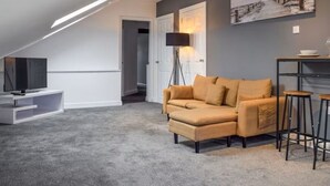 A modern living room with a grey carpet, a beige sofa, a TV on a white stand, a floor lamp, and a painting on the wall. - Elysian Residence (Newsome)