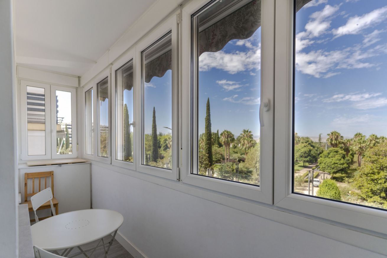 Een kamer met een witte tafel en stoel, grote ramen met decoratieve luiken, en uitzicht op bomen en een heldere lucht.